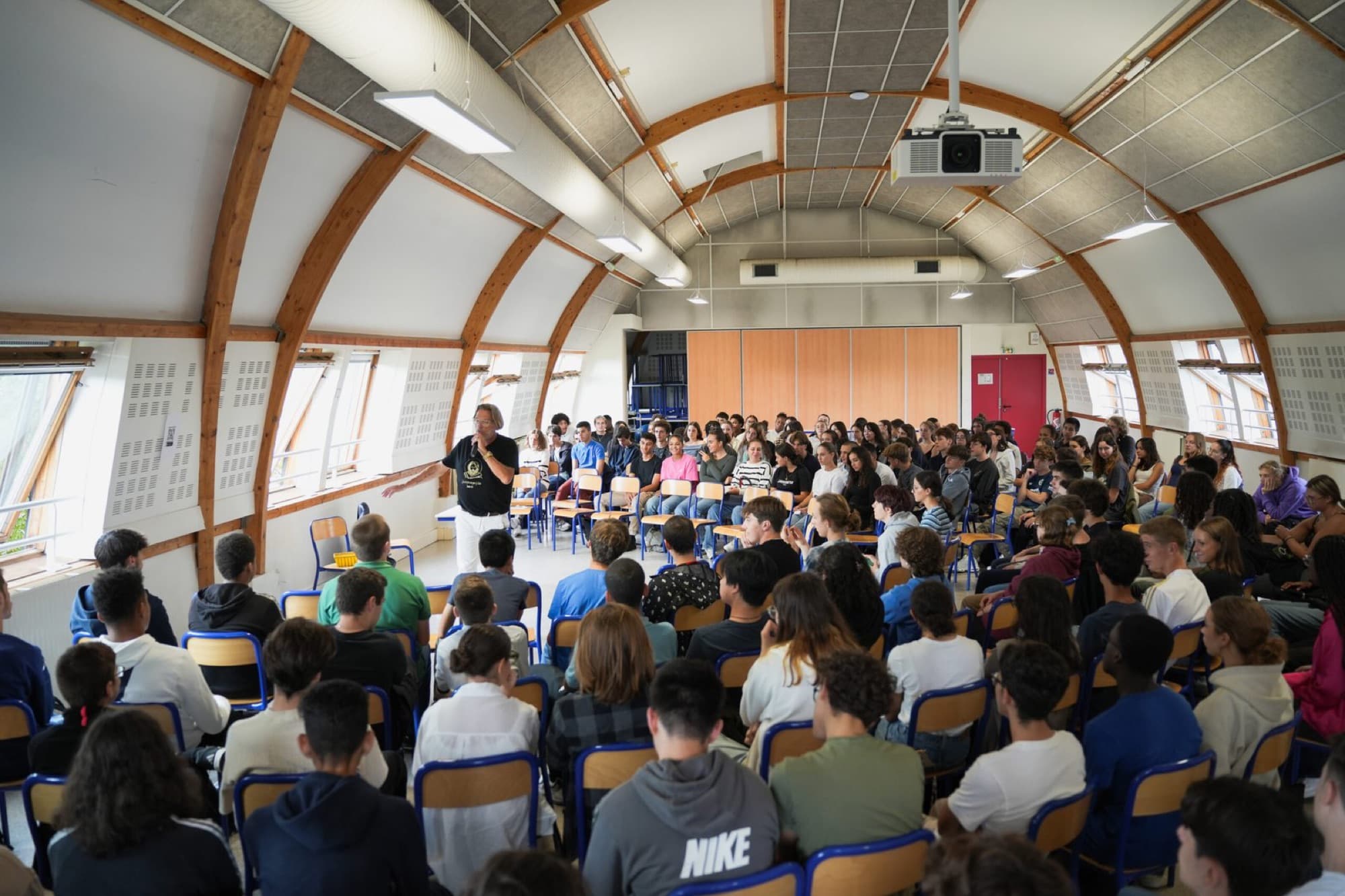 Une salle d'établissement scolaire remplie de lycéens assis face à un intervenant d'AALG qui prend la parole au micro.
